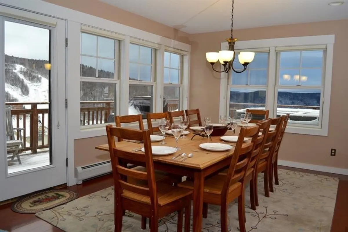 A large wooden dining table under a chandelier is framed by windows revealing scenic views, connecting the kitchen and dining experience to the outdoors. The setup evokes memorable meals and celebrations with loved ones.