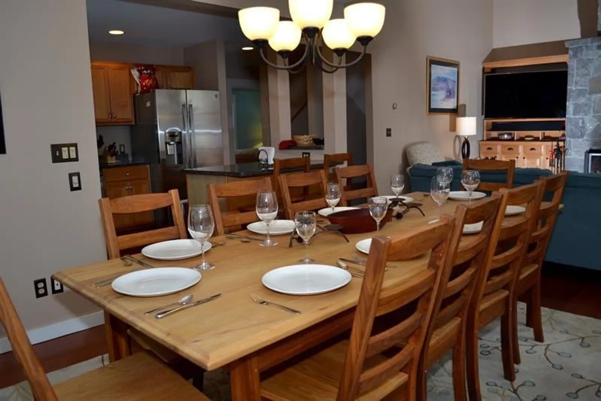A spacious and inviting dining area featuring a large wooden dining table under a statement chandelier. The background reveals an open kitchen and a comfortable seating area, all bathed in warm, welcoming light that suggests the perfect setting for group gatherings post-skiing.