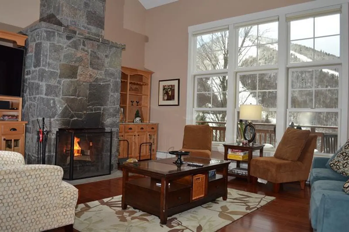 The image showcases a cozy living area with a striking stone fireplace as the centerpiece, surrounded by plush, inviting sofas. Large windows frame sweeping views of snow-dusted trees and mountains, filling the space with natural light. A coffee table sits ready for board games or morning coffee, creating an atmosphere perfect for relaxation and togetherness after a day on the slopes.