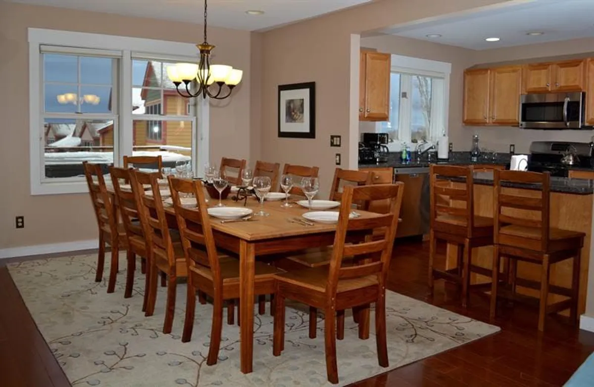 A spacious dining area with a large table, elegant chandelier, kitchen area in the background, and a prominent window view. The natural light streaming in hints at a picturesque setting just outside.