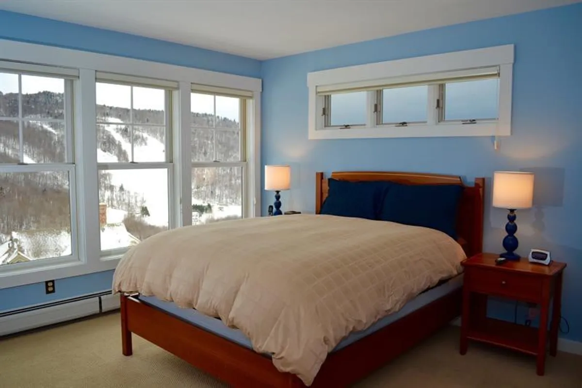 A serene bedroom featuring a bed with blue and beige bedding, a nightstand with a lamp, and large windows that frame a mountain view. The space feels peaceful and spacious, suggesting a restful retreat.