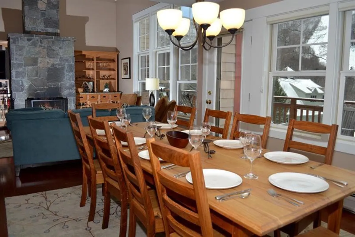 This scene shows a large dining table under a stylish chandelier, adjacent to a fireplace and surrounded by expansive windows that frame scenic mountain views. The arrangement suggests a seamless flow between dining and lounging areas, perfect for group meals and celebrations.