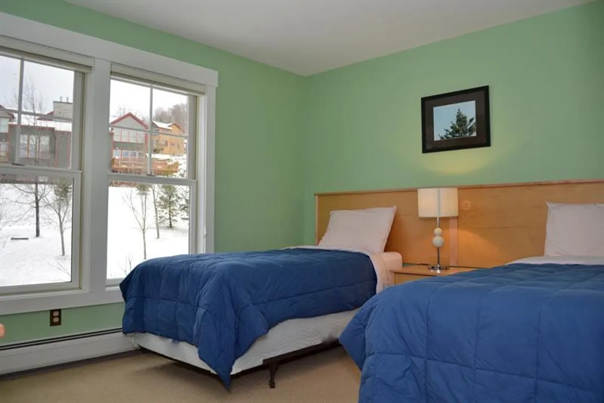 A cozy twin bedroom with two beds dressed in blue comforters, a large window letting in natural light, and framed artwork on the wall. The room feels tranquil and ideal for flexible sleeping arrangements.